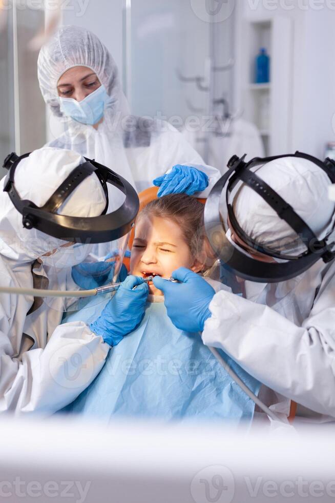 Mother comforting ittle girl with painfull face expression in the course of dental treatment. Stomatology team wearing ppe suit during covid19 doing procedure on child teeth. photo