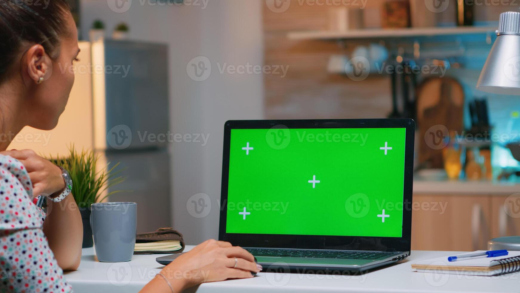 Freelancer looking at green screen monitor sitting at home in kitchen. Business woman watching desktop laptop display with green mockup, chroma key, during night time working overtime. photo