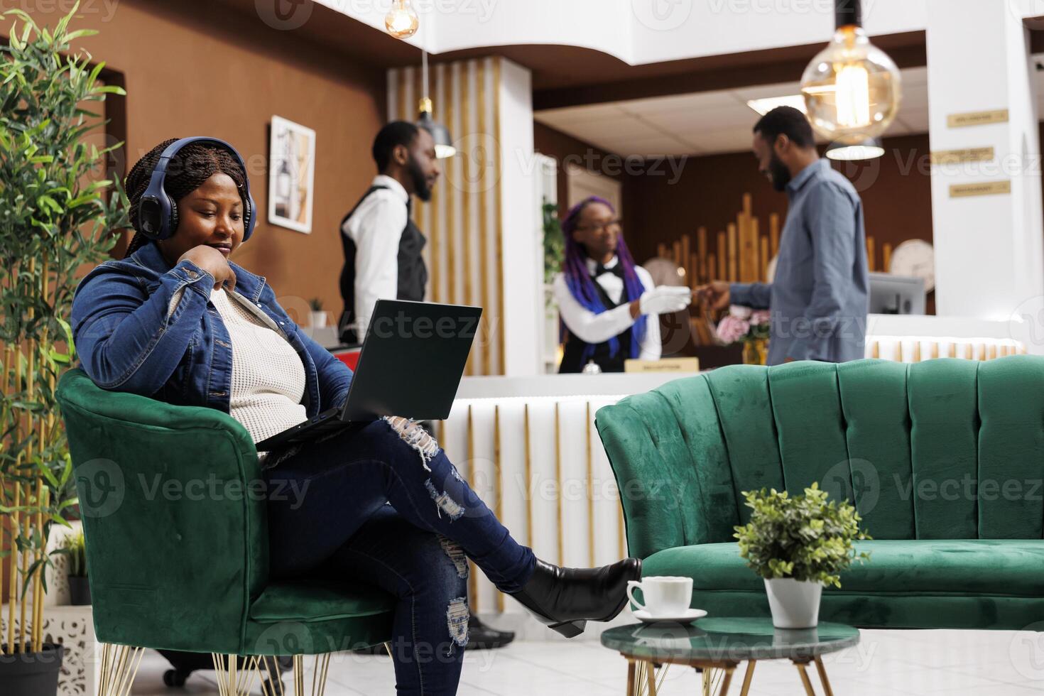 Young African American woman traveler sitting with laptop in hotel lobby using free wireless internet, watching movie online while waiting for check-in. Female freelancer working remotely from resort photo