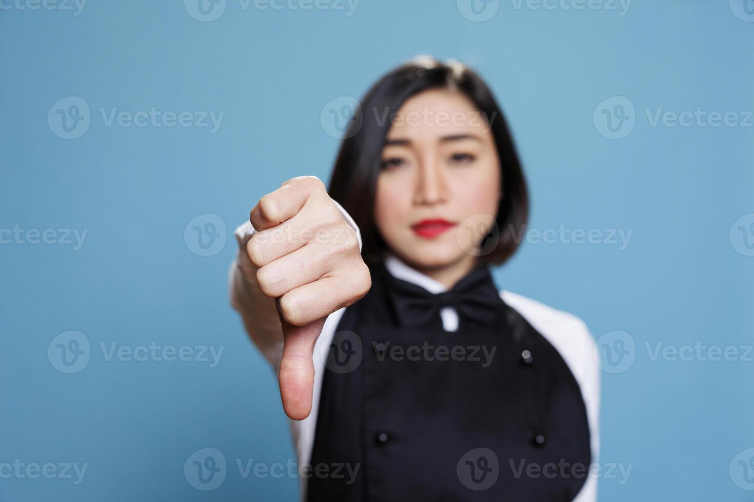 Young asian woman wearing waitress uniform showcasing disapproval with thumb down gesture portrait. Restaurant receptionist showing service negative feedback concept and looking at camera photo
