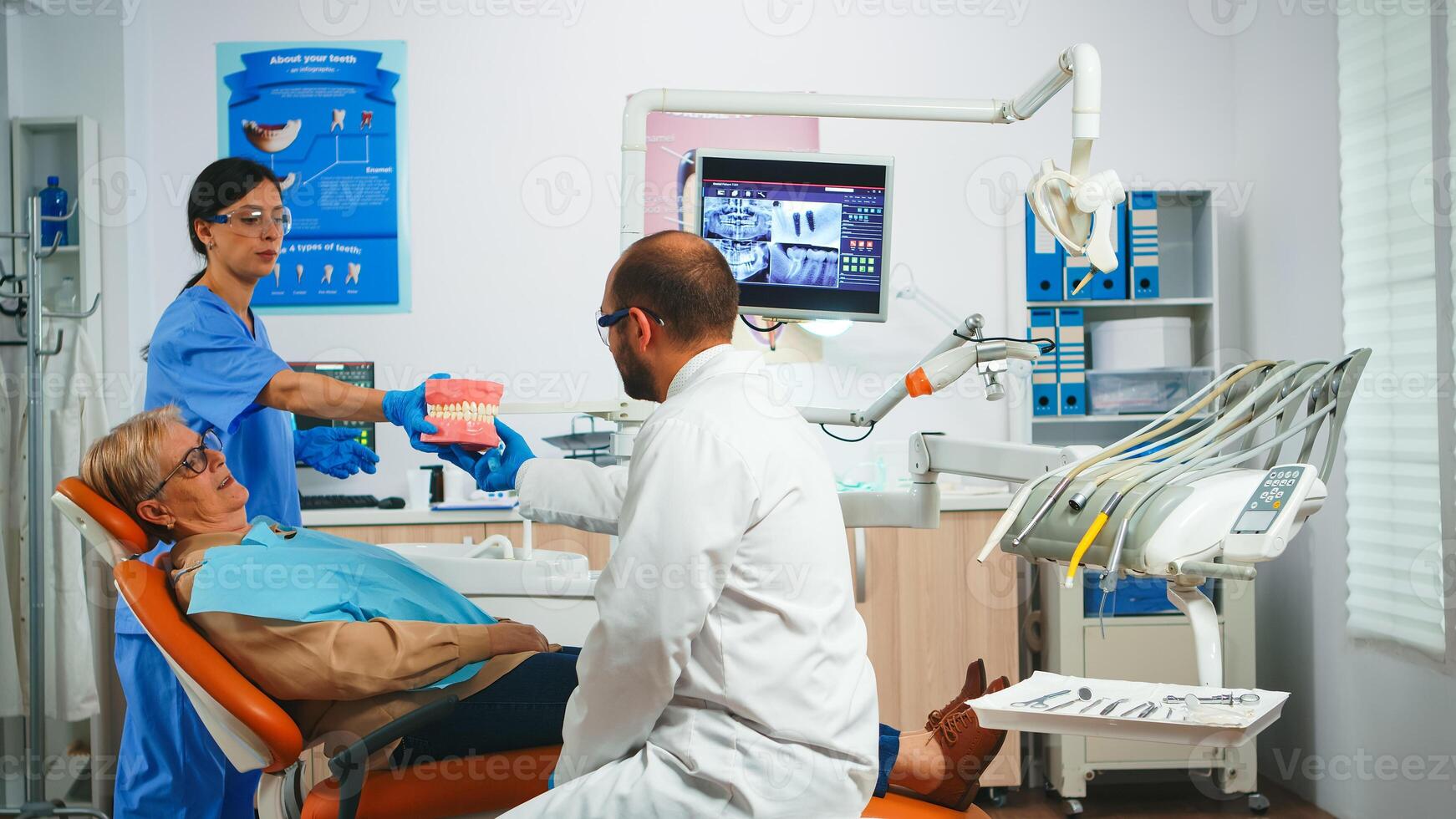 Stomatologist explaining proper dental hygiene using presentation teeth skeleton. Dentist telling to senior patient procedure holding sample of human jaw with a toothbrush in stomatology office. photo