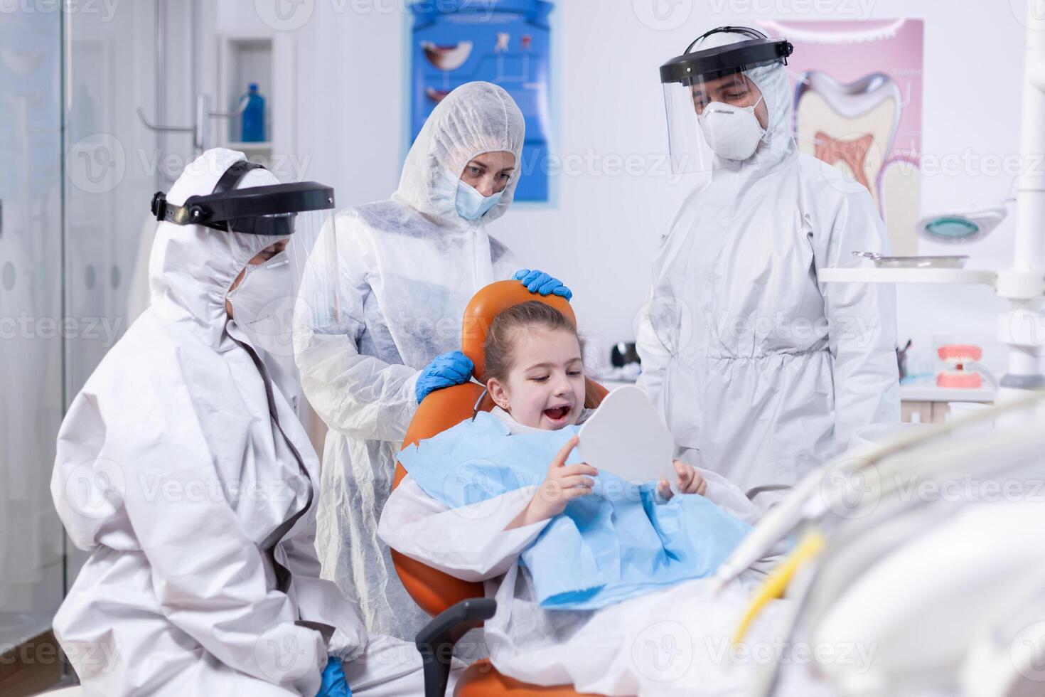 Little girl using dental mirror after mouth hygine procedure in the course of coronavirus pandemic. Child wearing ppe suit during teeth intervention at dental hospital. photo