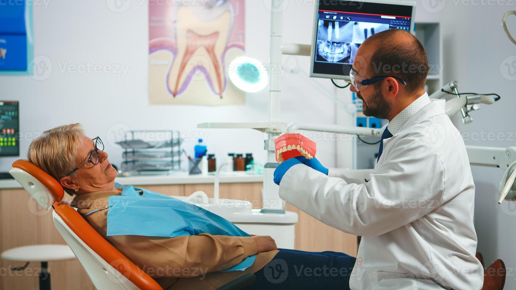 Doctor showing the correct dental hygiene using mock-up of skeleton of teeth. Dentist explaining proper dental hygiene to old patient holding sample of human jaw with toothbrush in stomatology office. photo