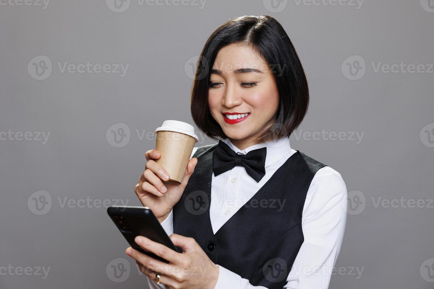 Smiling asian woman dressed in waitress uniform holding coffee paper mug and browsing internet on smartphone. Receptionist enjoying tea and engaging with social media networks on mobiel phone photo