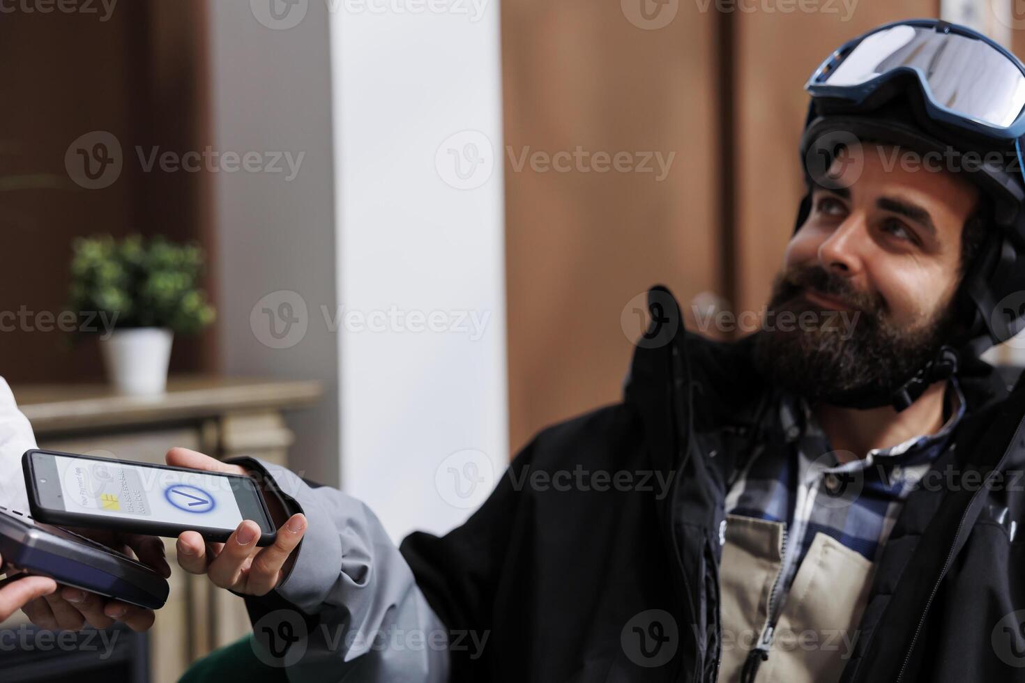 In ski resort lounge area, caucasian man pays with cell phone while staff assists with pos machine. Close-up view of male traveler wearing wintersport outfit using smart device for nfc payment. photo