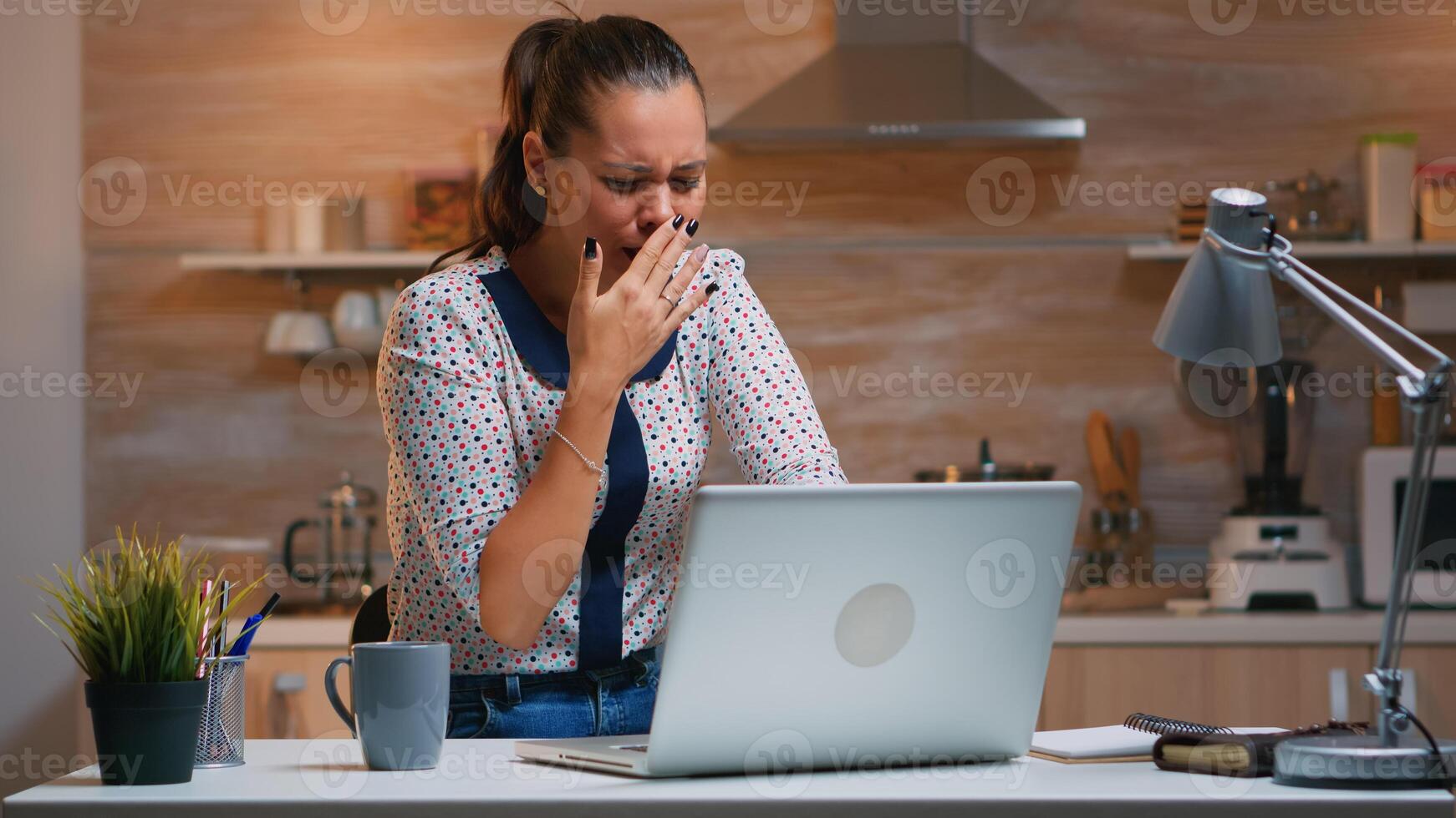 Woman yawning while working remotly from home late at night in the kitchen. Busy exhausted employee using modern technology network wireless doing overtime for job reading typing, searching photo