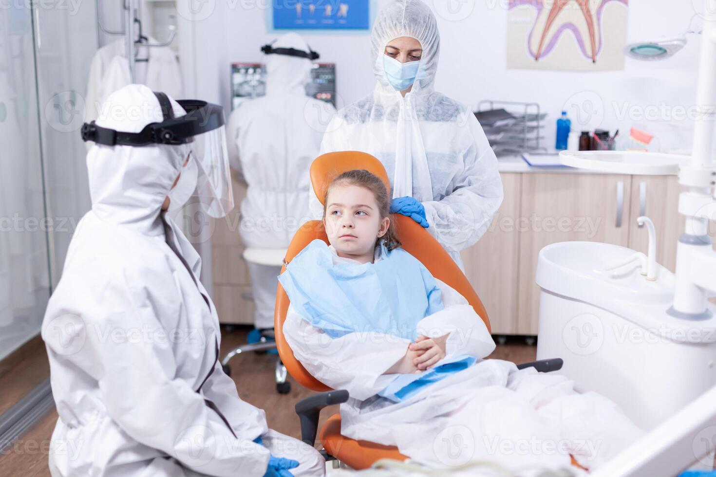 Serious little girl sitting in dental chair listening dentist talking about treatment for cavity. Stomatologist during covid19 wearing ppe suit doing teeth procedure of child sitting on chair. photo