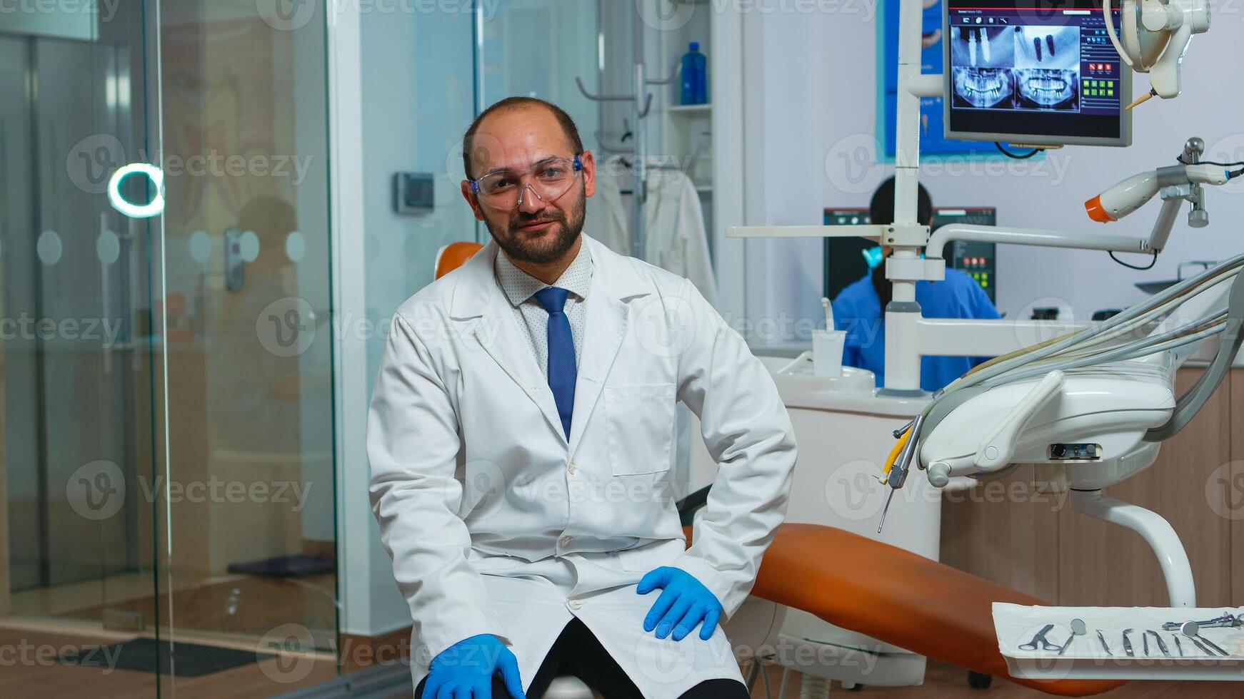 Dentist doctor looking at camera talking with patients about dental hygiene. Stomatologist speaking on webcam sitting on chair in stomatological clinic with assistent in background working on computer photo