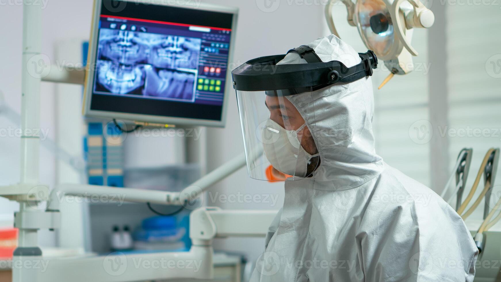Portrait of dentist with protection mask speaking with elderly patient during global pandemic. Orthodontic doctor wearing protective suit, coverall, face shield and gloves in coronavirus period. photo