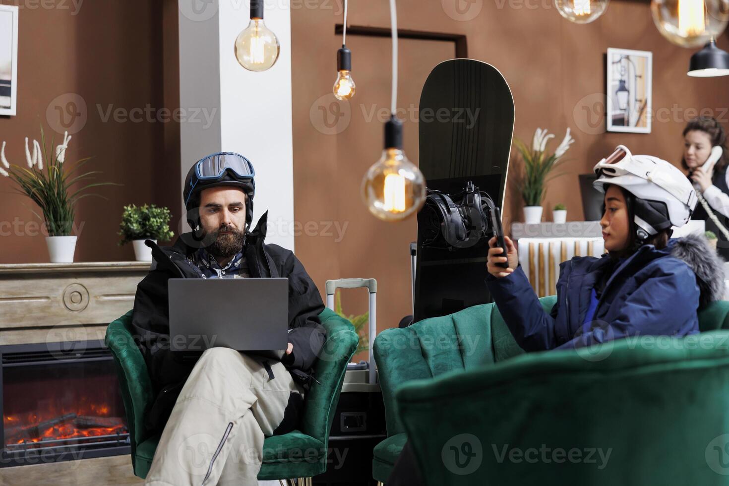 Two travelers in snow clothing using their technology devices while waiting for check-in process at luxury winter resort. Couple holding their laptop and phone while seated in ski hotel reception. photo