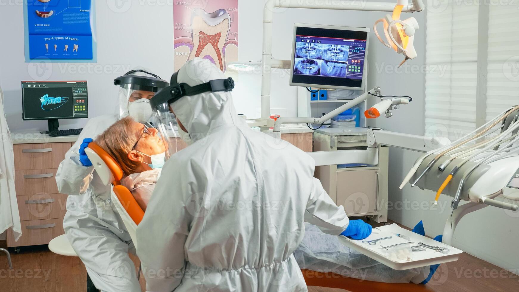 Nurse and doctor in protection suit working in dental unit during coronavirus pandemic treating senior patient. Assistant and orthodontic doctor wearing coverall, face shield, mask and gloves. photo