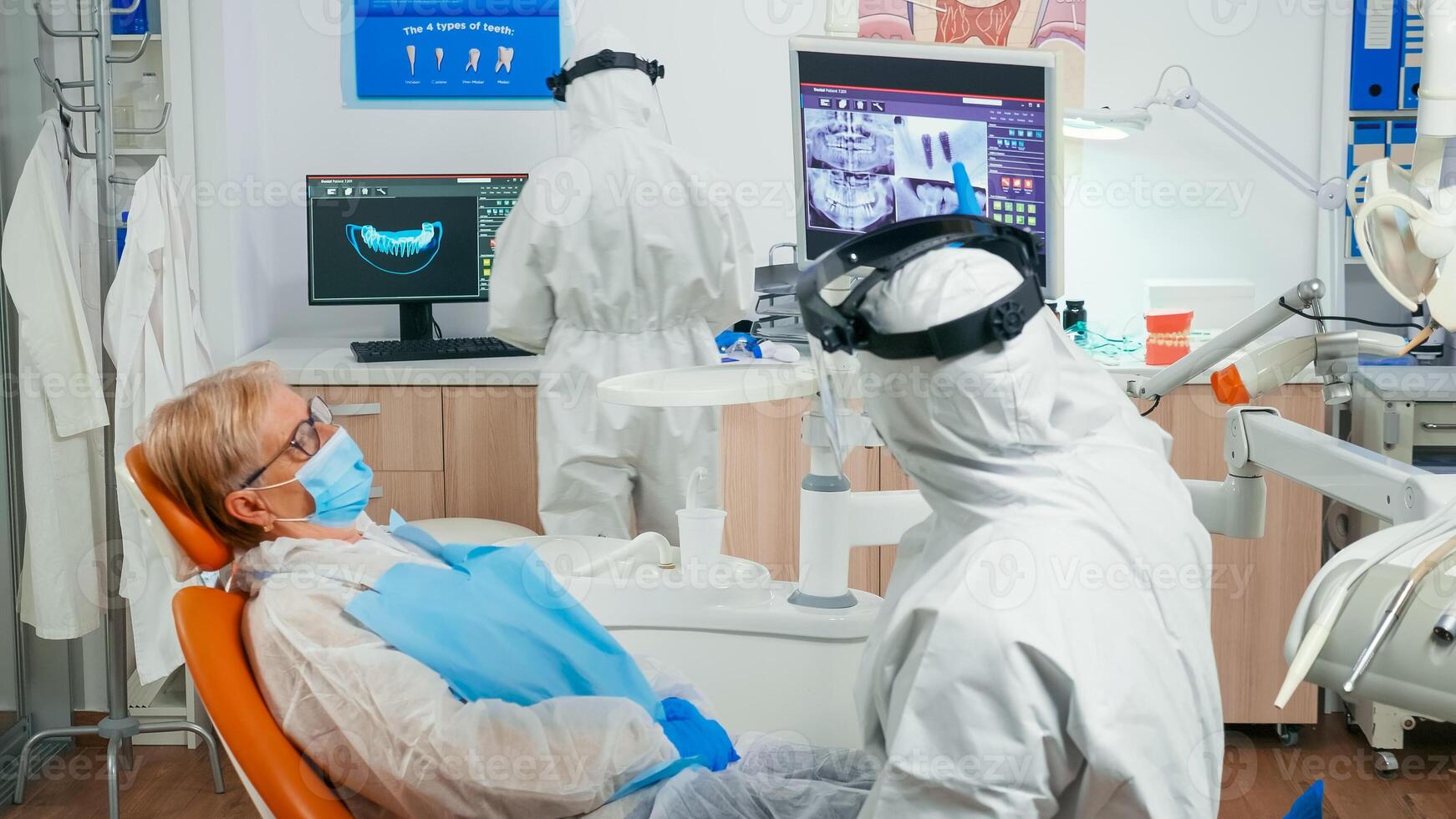 Stomatologist in coverall and patient looking at digital dental x-ray reviewing it before surgery during covid-19 pandemic. Medical team wearing face shield, protection suit mask and gloves in clinic photo