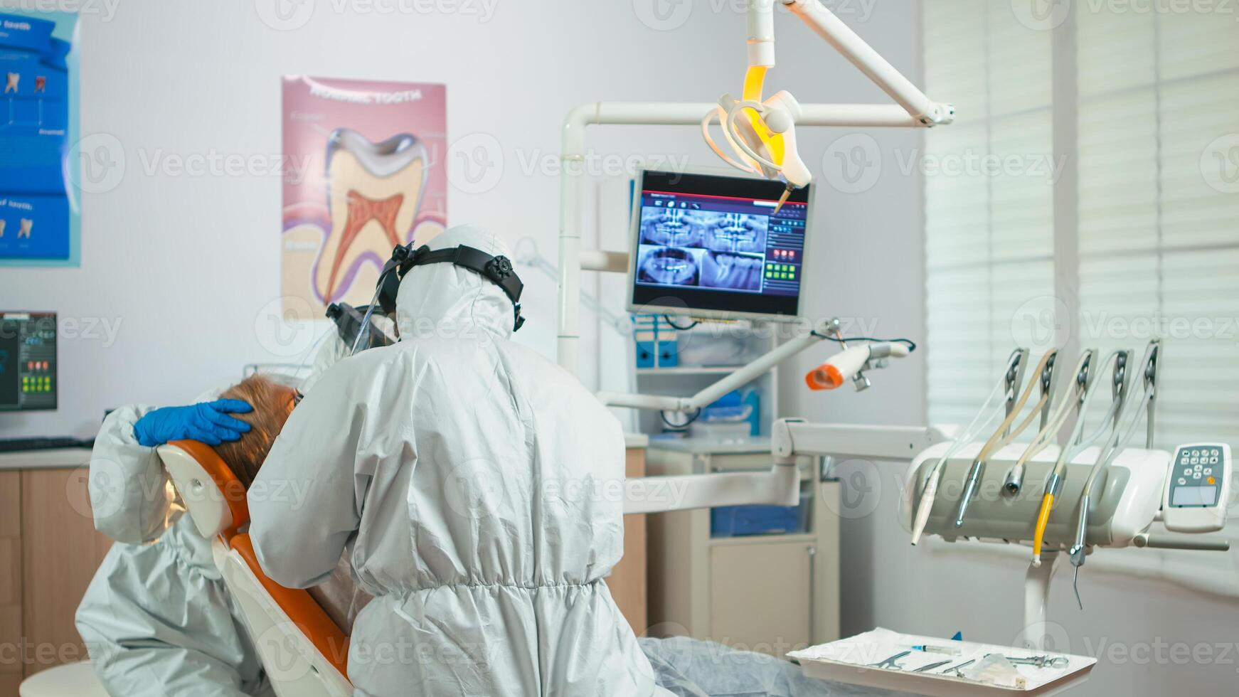 Doctor in protection suit using drilling machine during dental examining in pandemic period. Orthodontist and nurse working wearing coverall, face shield, mask, gloves in stomatological office photo