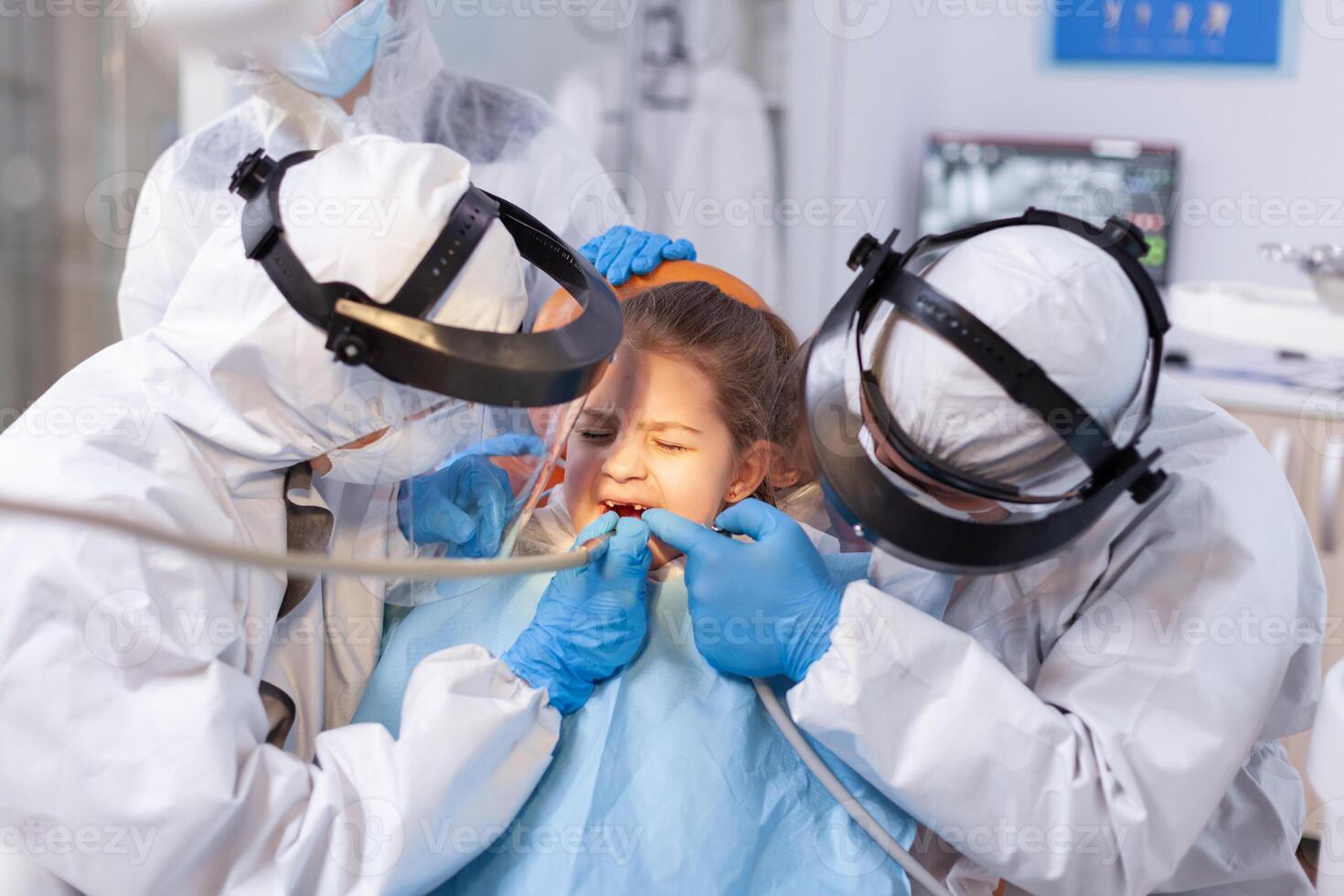 Woman dentist doctor treating little girl tooth with pain face expression. Stomatology team wearing ppe suit during covid19 doing procedure on child teeth. photo