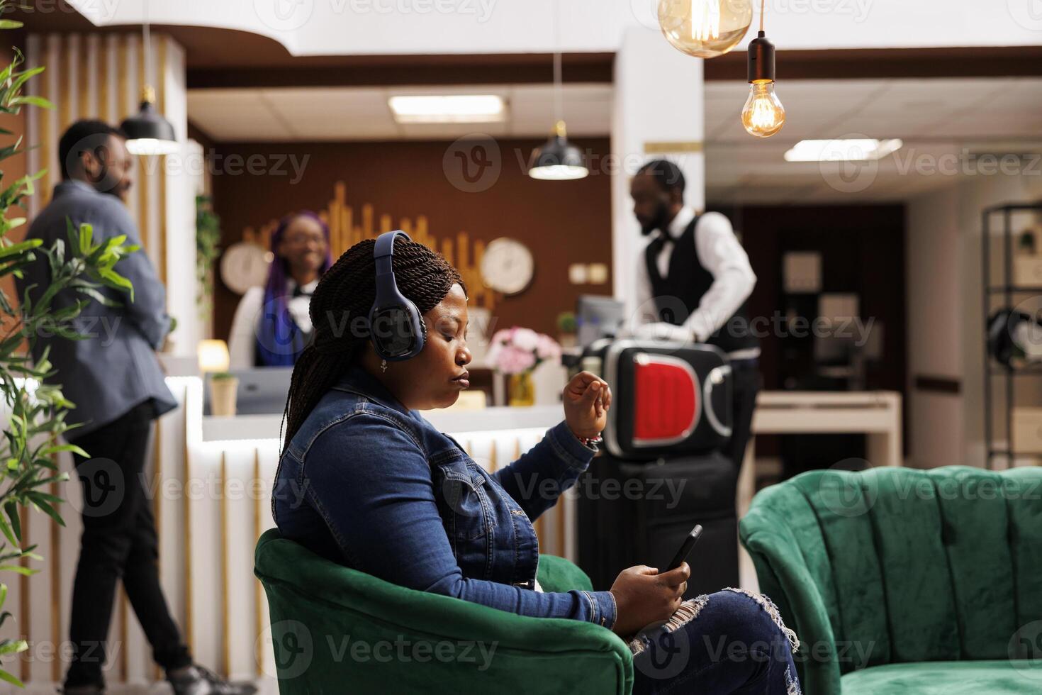 Serious African American woman tourist wearing headphones using smartphone while sitting at hotel lounge area against front desk. Focused female guest completing check-in process on mobile device photo
