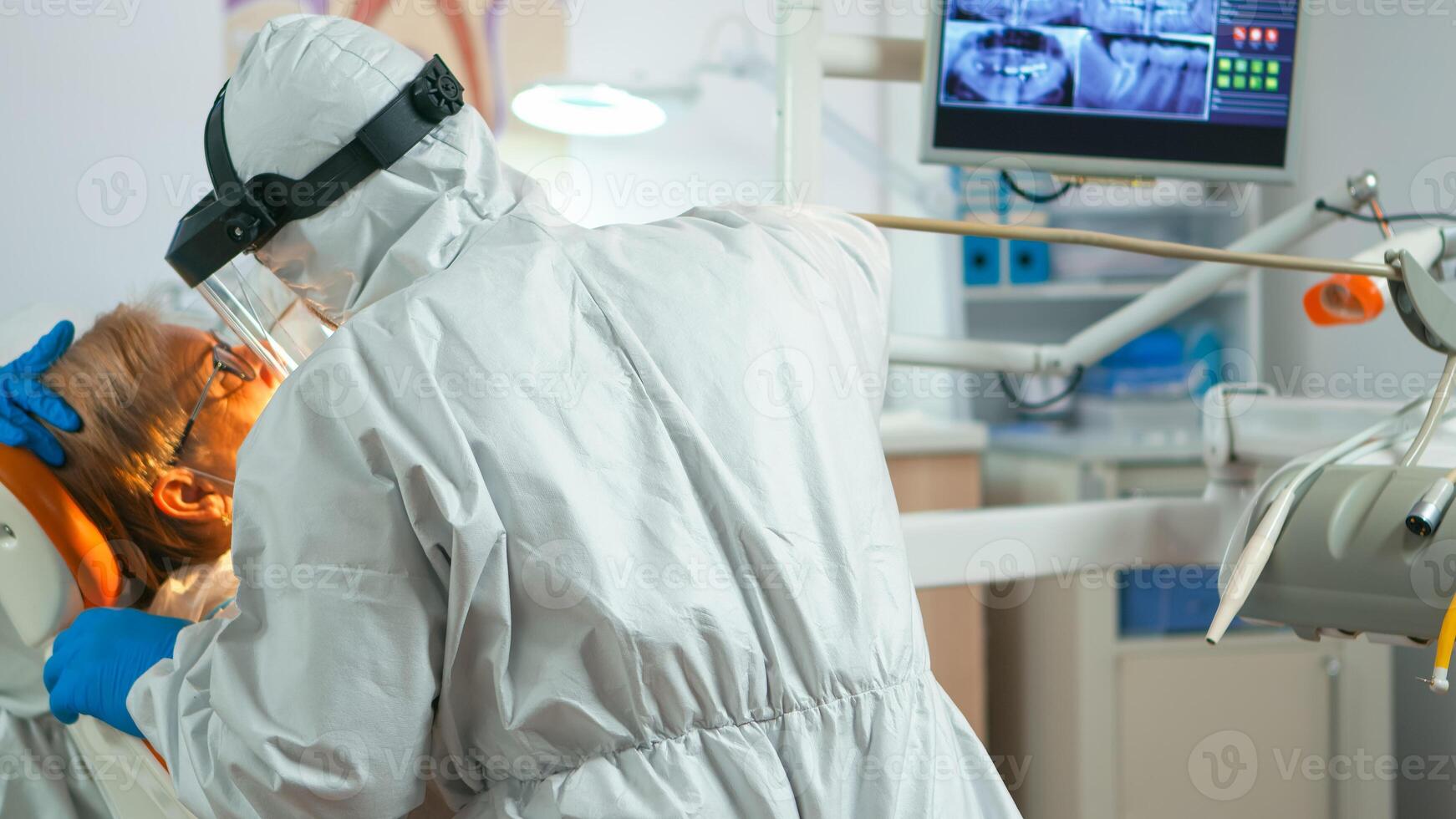 Close up of dentistry doctor in coverall using drilling machine for examining patient during global pandemic. Medical team wearing protective suit,, face shield, mask, gloves in stomatological office photo