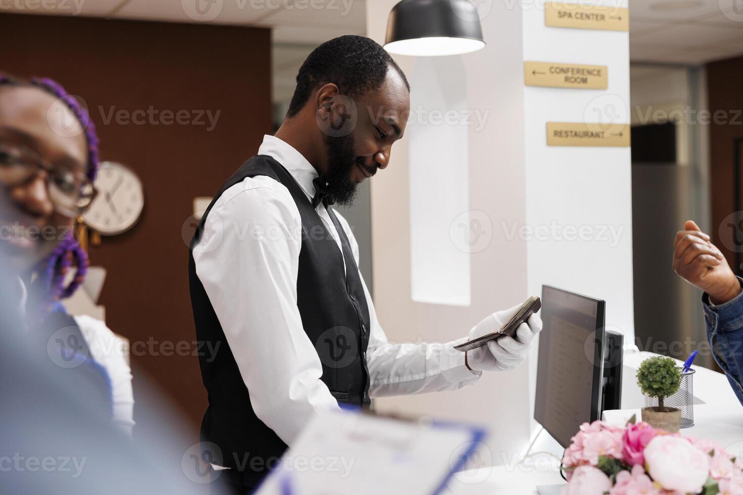 Smiling friendly African American man receptionist checking tourist passport at hotel check-in. Positive front desk staff registering guests at reception counter, completing registration procedure photo