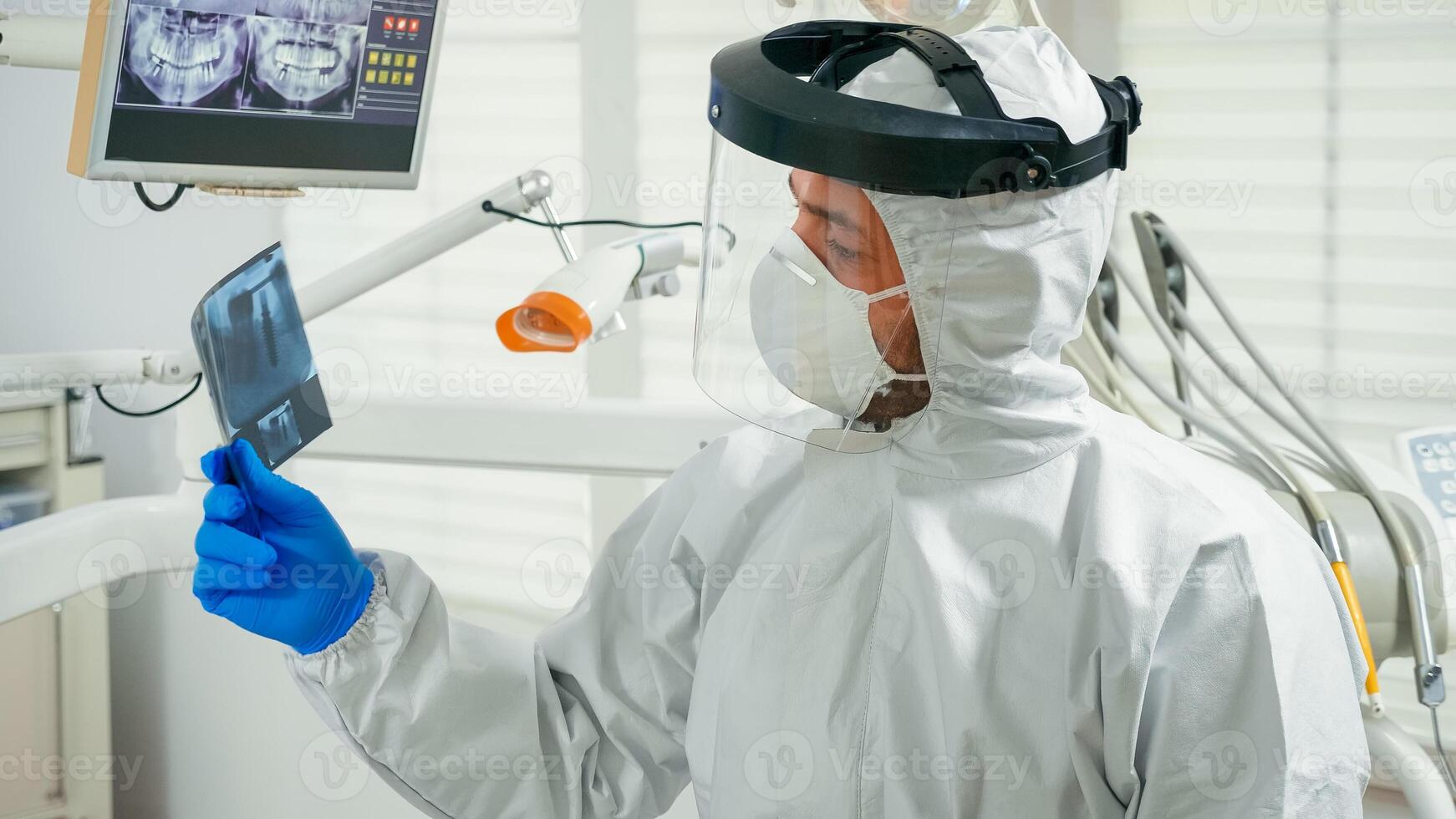 Portrait of dentist with face shield explaining holding dental x-ray to patient pointing on monitor during covid-19 pandemic. Orthodontic doctor wearing protection suit, coverall, mask and gloves photo