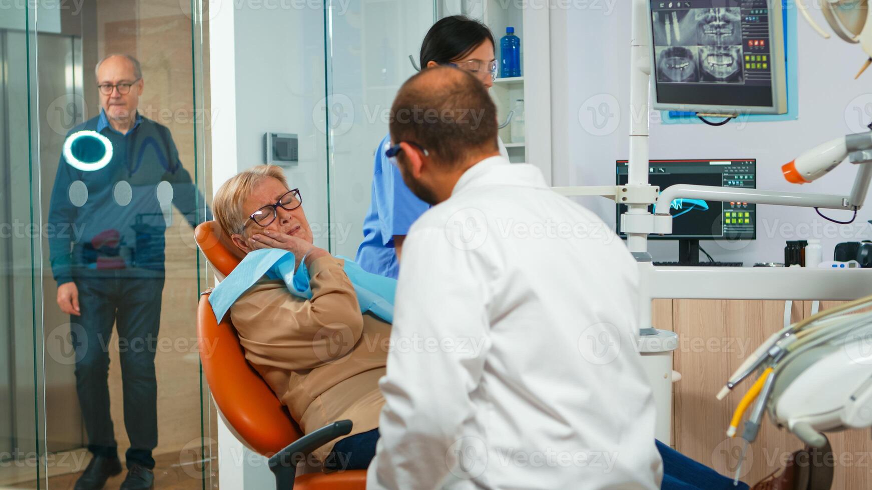 Woman with toothache talking with orthodontic showing affected mass. Elderly patient explaining dental problem to doctor while sitting on stomatological in modern private clinic before intervetion photo