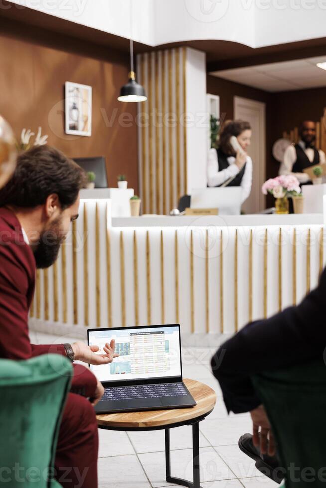 Employees meeting at hotel reception to work on collaboration, forming international connections in navigating global business landscape. Entrepreneurs reading shared ideas on laptop. photo