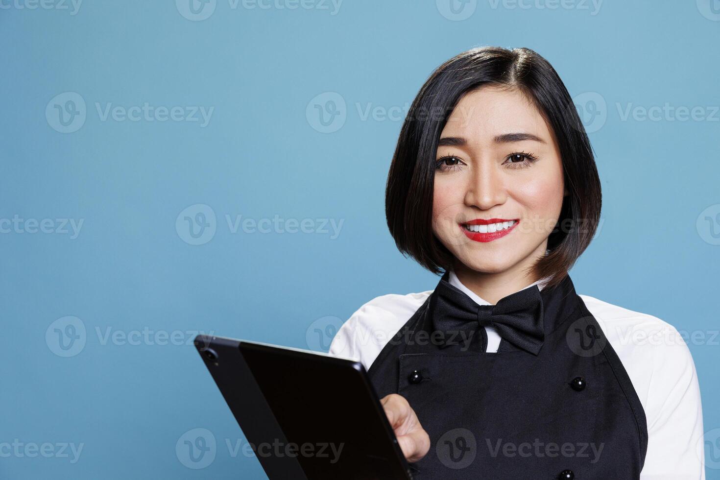 Smiling cafe waitress holding digital tablet at work and looking at camera with cheerful facial expression. Joyful cafe woman worker using portable gadget portrait on blue background photo