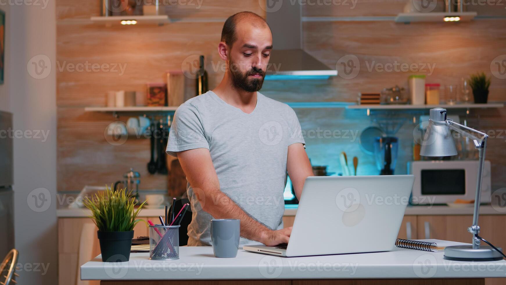 Exhausted freelancer yawning sitting at kitchen desk in front of laptop late at night. Busy focused employee using modern technology network wireless doing overtime for job reading writing, searching photo