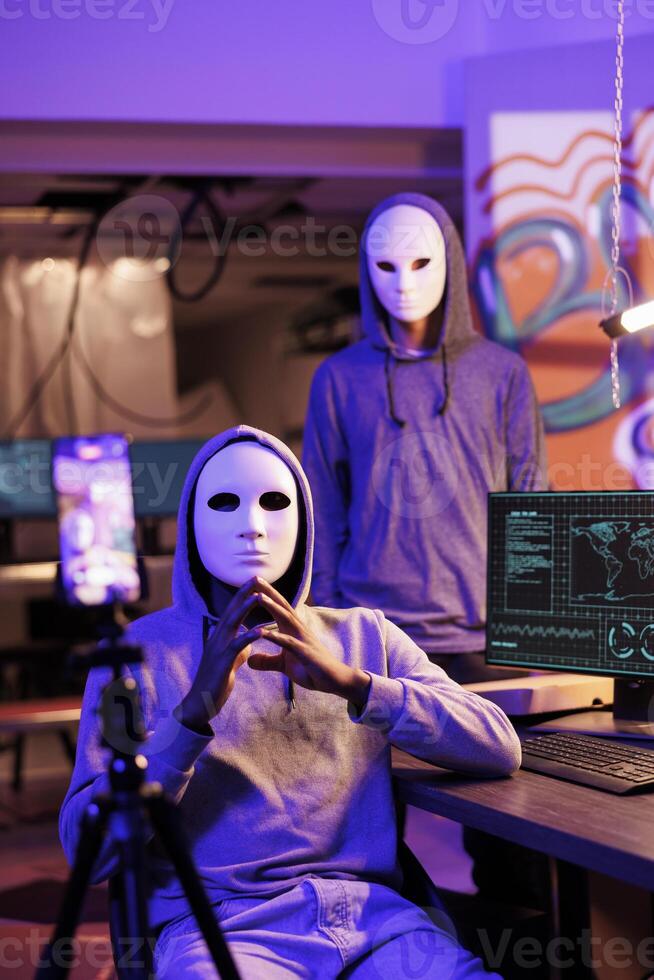 Scammers in anonymous masks streaming threat online and demand victim for payment. Hackers with hidden identity filming ransom message on mobile phone in abandoned warehouse photo