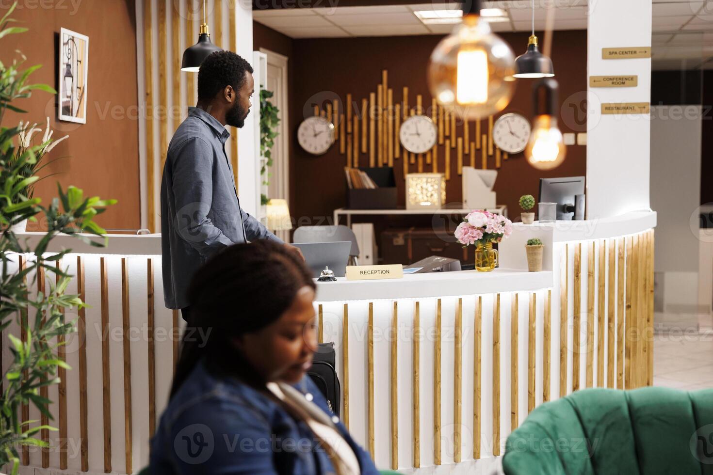 Young African American man tourist waiting for receptionist in reception area, arriving at hotel. Black guy traveler with suitcase standing at empty front desk, wait for check in procedure photo