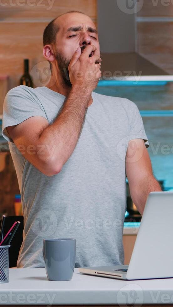 Exhausted freelancer yawning sitting at kitchen desk in front of laptop late at night. Busy focused employee using modern technology network wireless doing overtime for job reading writing, searching photo