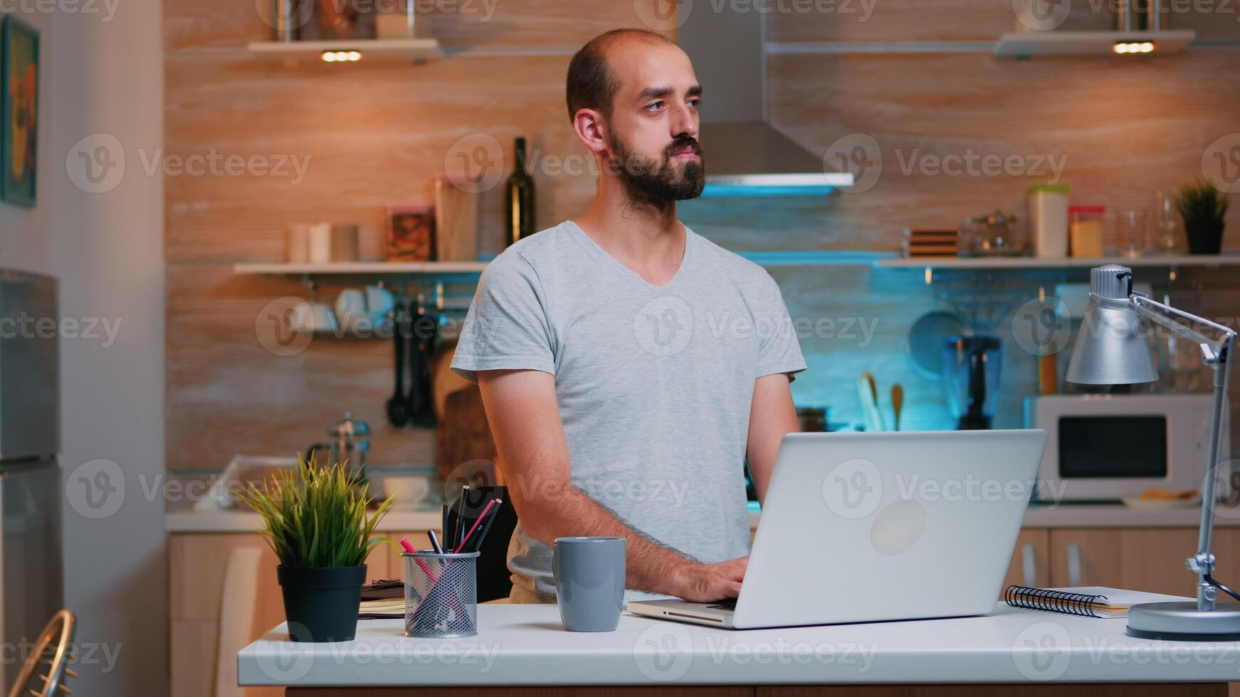Man thinking at next project while working from home reading the tasks looking on laptop sitting in home kitchen. Busy employee using modern technology network wireless doing overtime reading writing, searching photo