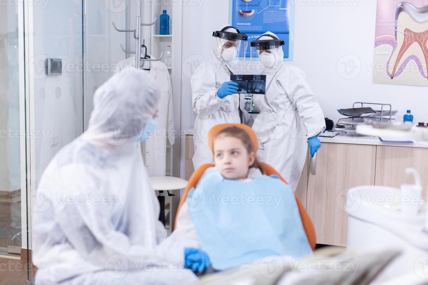 Dentist team analyzing kid radiography dressed in protection gear against coronavirus. Stomatologist during covid19 wearing ppe suit doing teeth procedure of child sitting on chair. photo