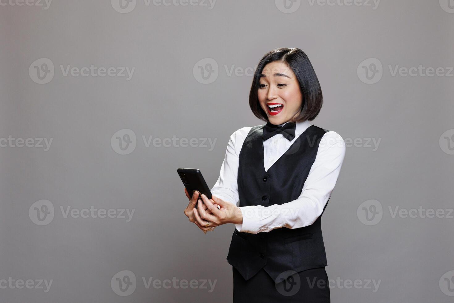 Excited happy asian waitress reading catering service positive reviews while holding smartphone. Young woman hotel receptionist in uniform checking good news message on mobile phone photo