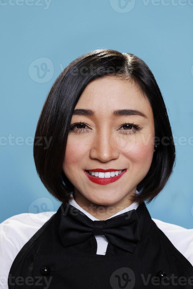 Smiling young attractive asian woman wearing waitress black and white uniform with bow tie closeup portrait. Cheerful woman receptionist looking at camera with positive facial expression photo