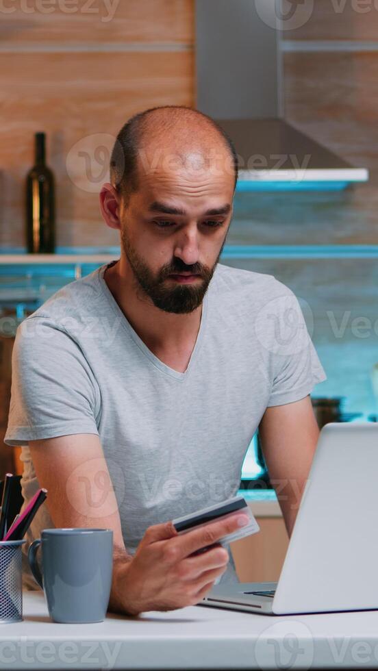 Young man shopping online late at night sitting in the kitchen on laptop using credit card. Employee making paymant transaction from home on digital notebook conected to internet photo