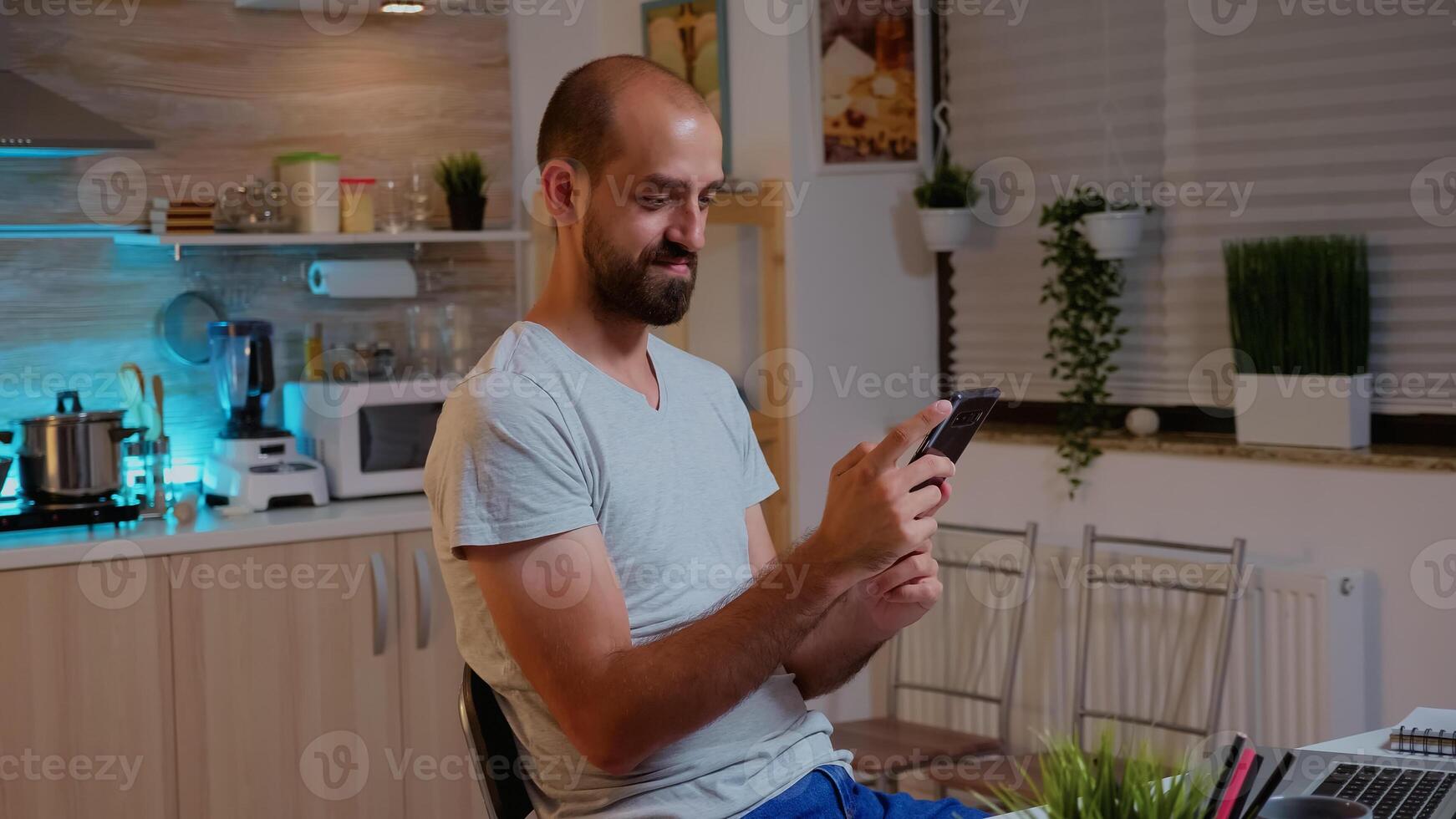 Freelancer man using phone and smile while working overtime from home. Busy focused employee using modern technology network wireless late at night reading writing, searching photo