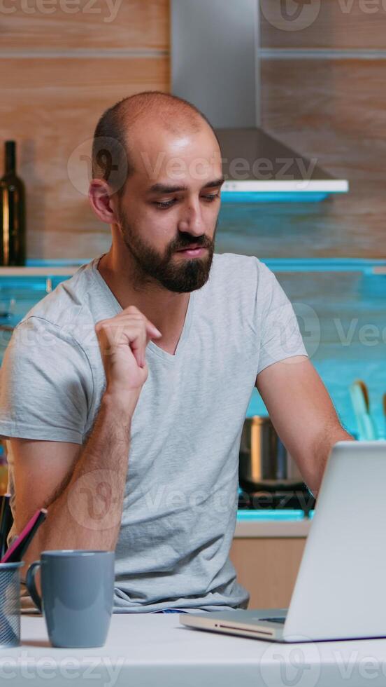 Remote man entrepreneur having video conference with colleagues using laptop sitting in the kitchen late at night. Busy employee using modern technology network wireless reading writing, searching photo