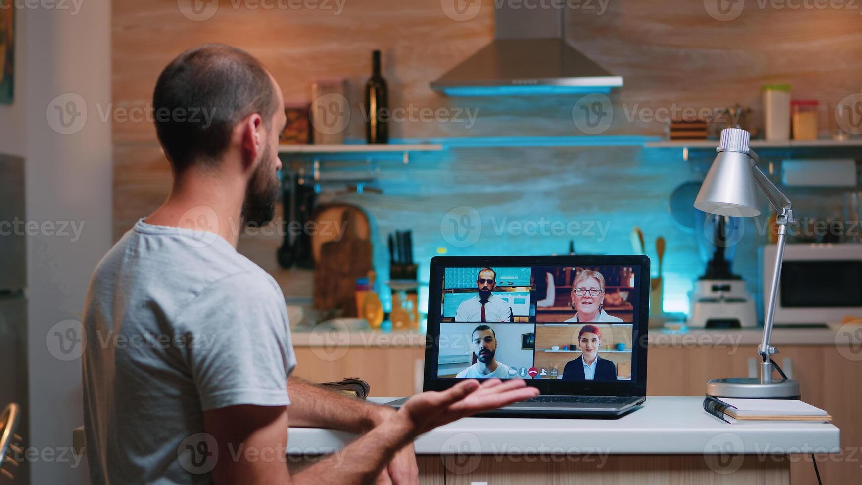 Employee working remotely discussing with partners online using laptop sitting in the kitchen. Freelancer using modern technology network wireless talking on virtual meeting at midnight doing overtime photo