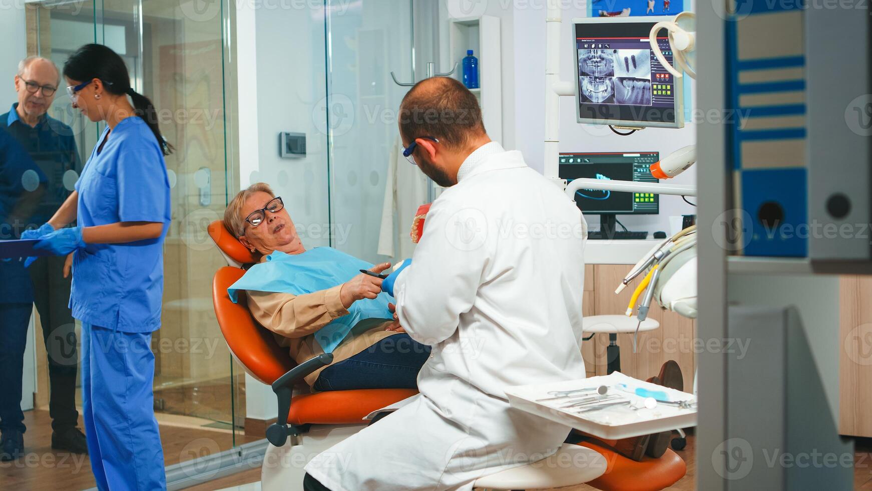 Young man dentist explaining the surgery using a plaster model of the mandible. Stomatologist holding mock-up of skeleton of teeth, sample of human jaw with toothbrush speaking with elderly patient. photo