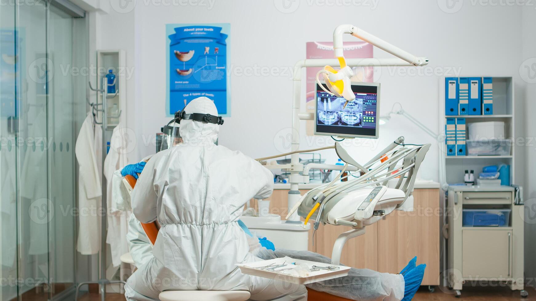 Dentist and assistant in coverall examining mouth of senior patient during global pandemic. Medical team working wearing overall, face shield, protection suit, mask, gloves in stomatological office photo