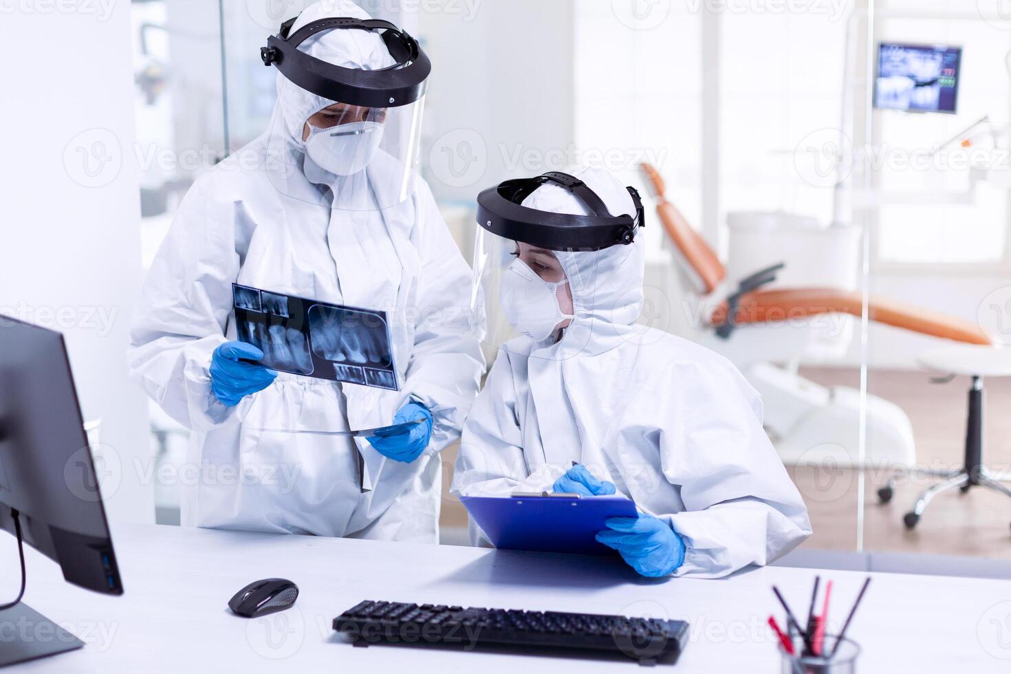 Stressed dentist and nurse examining teeth x-ray wearing ppe suit. Medical specialist wearing protective gear against coronavirus during global outbreak looking at radiography in dental office. photo