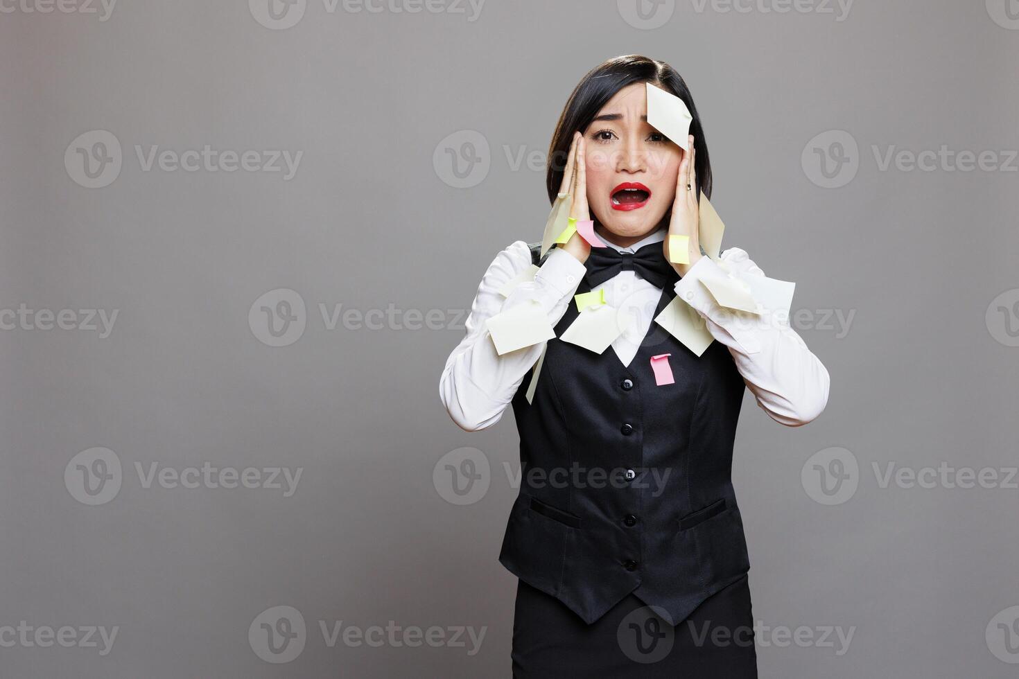 Exhausted busy restaurant manager in uniform overloaded with tasks panicking and looking at camera. Catering service stressed asian waitress with sticky notes on face portrait photo