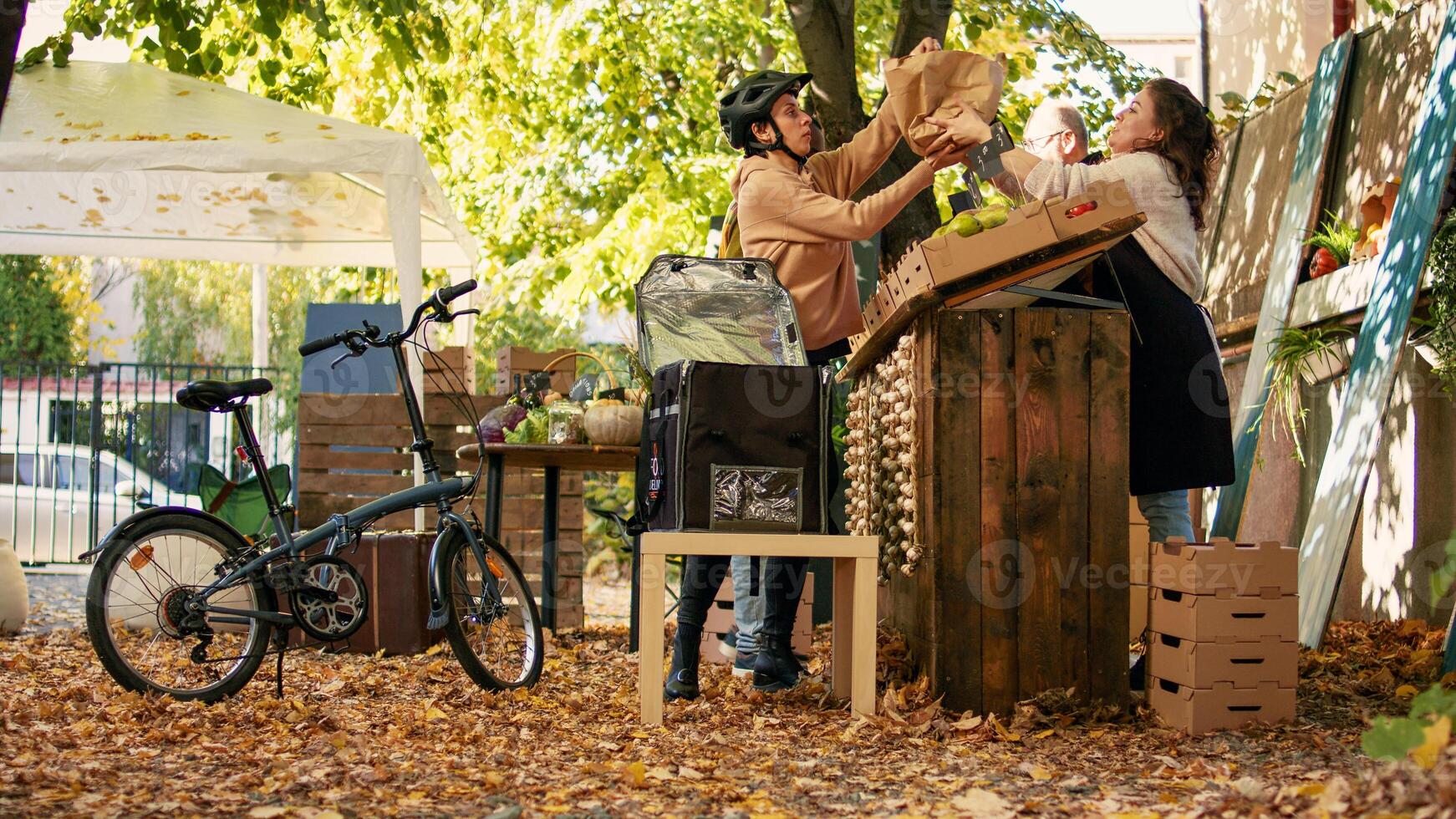 Female delivery employee with bike picking up food order at local marketplace, delivering natural products. Young courier standing near farm produce stand, taking eco fruits and vegetables. photo