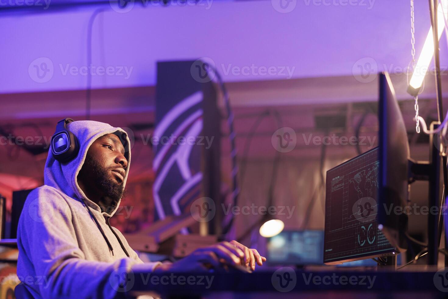 Relaxed african american criminal in hood hacking computer server while listening to music in headphones. Man hacker in earphones wearing hood planning phishing online scam photo