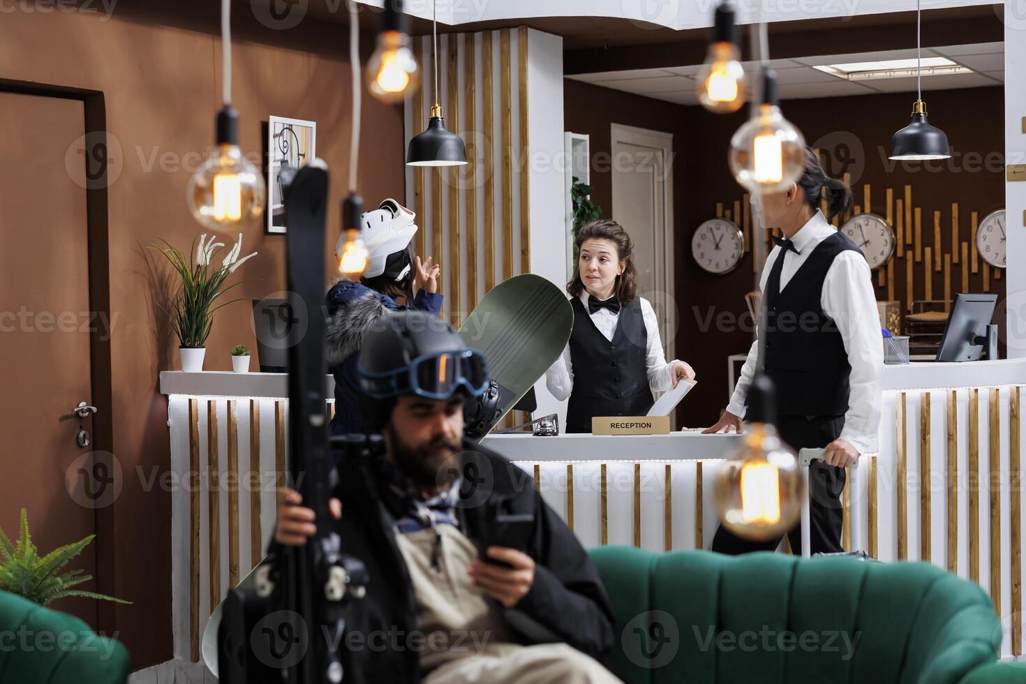 In hotel lobby, staff assist female tourist with check-in while male customer browses on cell phone. Winter travelers prepare for skiing and snowboarding while employees provide good customer service. photo