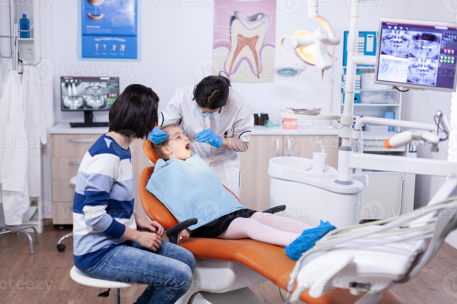 Kid patient in dentistiry office getting treatment for teeth cavity sitting on dental chair wearing bib. Dentistry specialist during child cavity consultation in stomatology office using modern technology. photo
