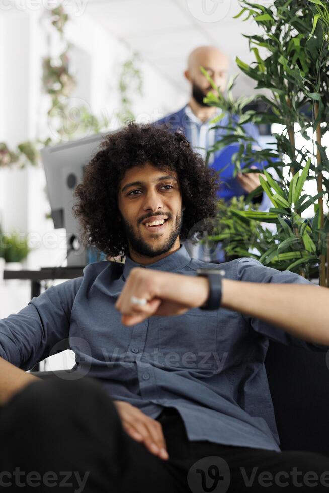 Cheerful businessman planning meeting schedule using smartwatch voice record app. Arab young man professional tracking activity on fitness trackers while working in business office photo