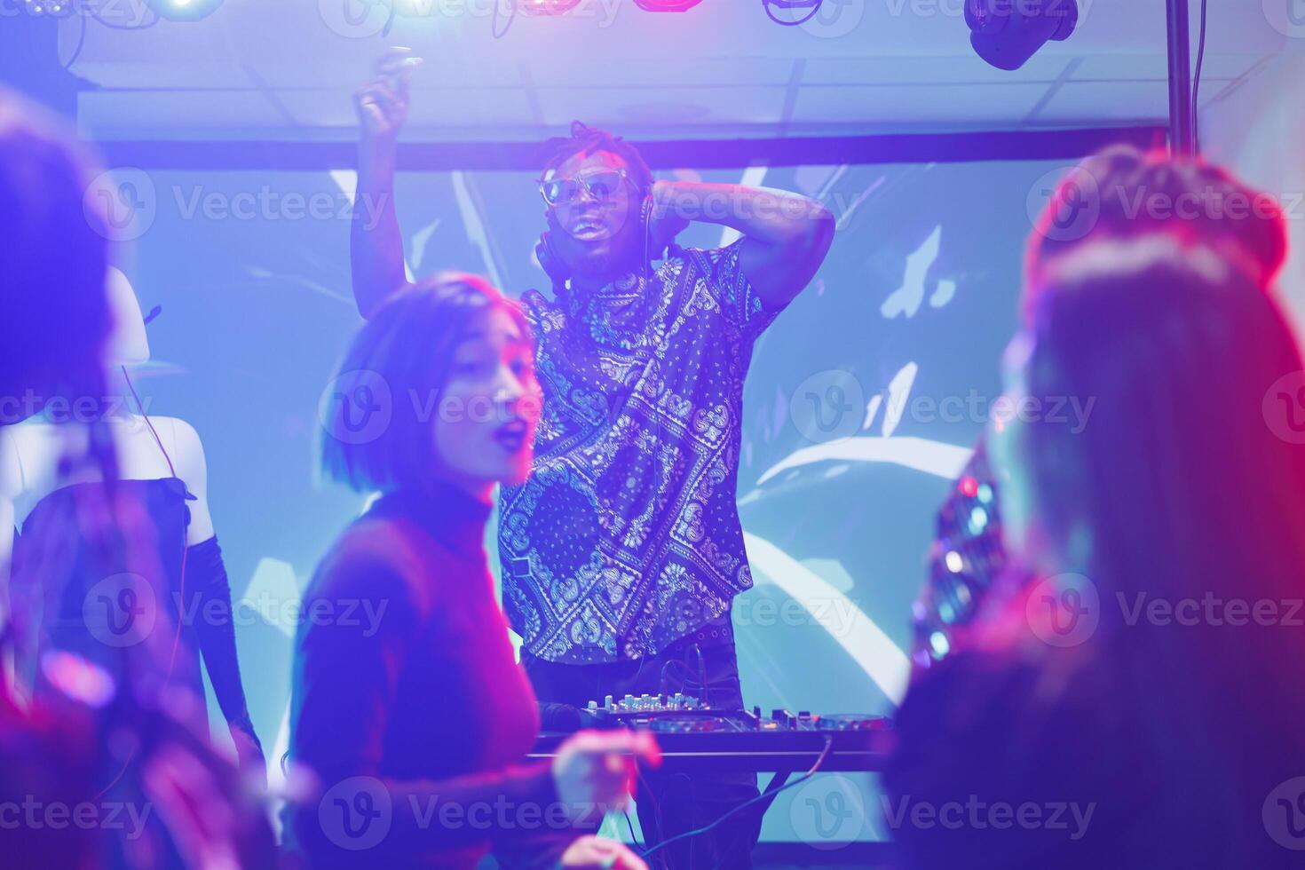 People partying on dancefloor while young dj performing on stage in nightclub. Diverse clubbers crowd dancing at discotheque while musician playing electronic music in club photo