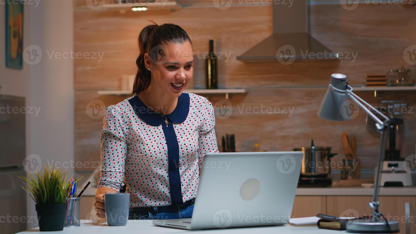 Woman remote entrepreneur having video conference with colleagues using laptop sitting in the kitchen late at night. Busy employee using modern technology network wireless doing overtime for job. photo