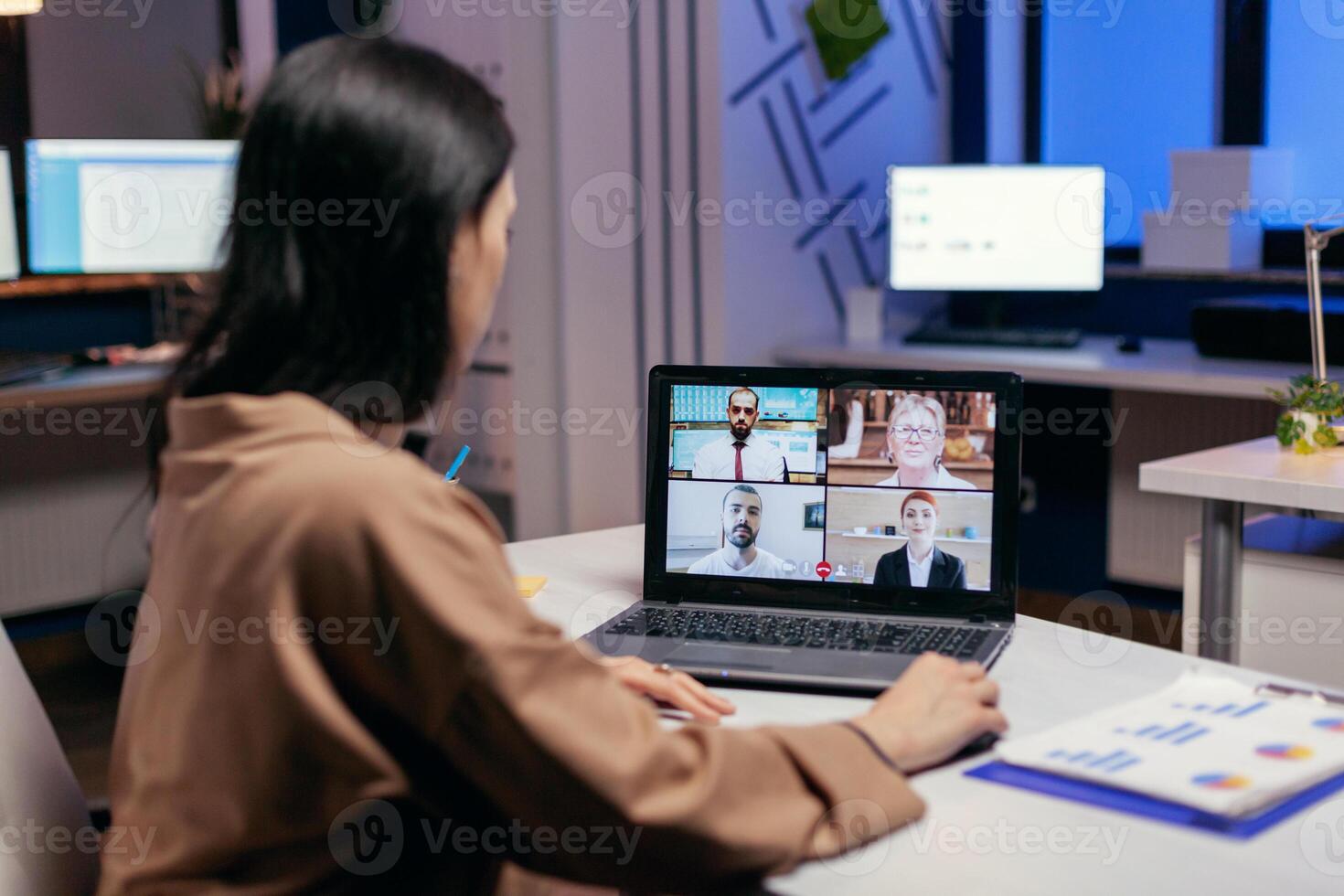 Entrepreneur talking in the course of online meeting with colleagues doing overtime. Woman working on finance during a video conference with coworkers at night hours in the office. photo