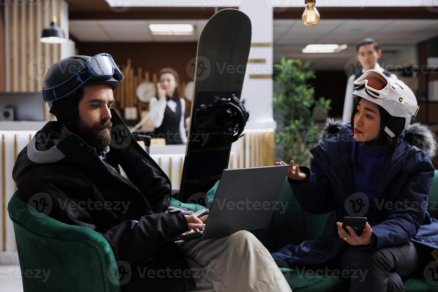 In hotel lobby young guests on couch plan skiing and snowboarding activities with digital devices. Couple using technology for online check-in at ski resort. Snowboard helmets and laptop available. photo